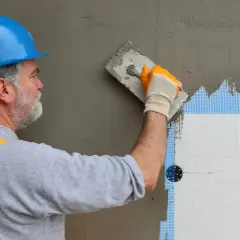 Pracownik nakłada klej na styropian, używając siatki zbrojącej. Cena robocizny zależy od jakości materiałów.