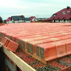 Budowa stropu ackermana. Czerwone pustaki ceramiczne ułożone w rzędach, gotowe do zalania betonem. W tle domy z czerwonymi dachami.