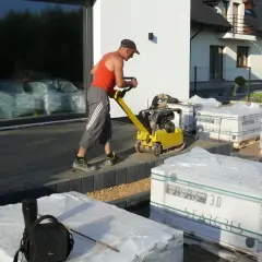Mężczyzna ubija podłoże zagęszczarką, przygotowując je pod **masę wyrównującą** i kostkę brukową.