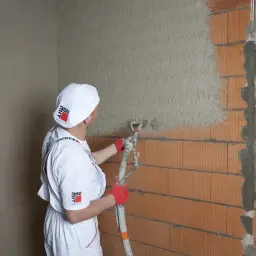 Pracownik w białym stroju nakłada tynki cementowo wapienne na ścianę z pustaków ceramicznych za pomocą agregatu.