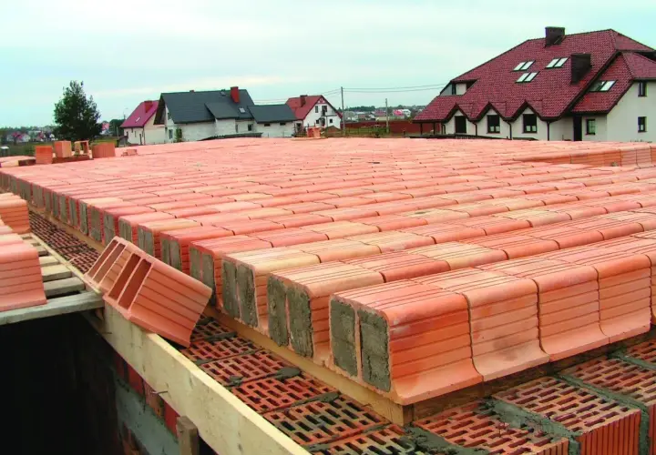 Budowa stropu ackermana. Czerwone pustaki ceramiczne ułożone w rzędach, gotowe do zalania betonem. W tle domy z czerwonymi dachami.
