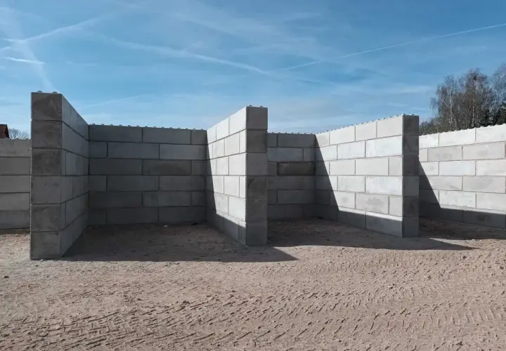 Budowa muru oporowego z bloczków betonowych. Konstrukcja w kształcie litery L, z widocznymi cieniami na piasku.