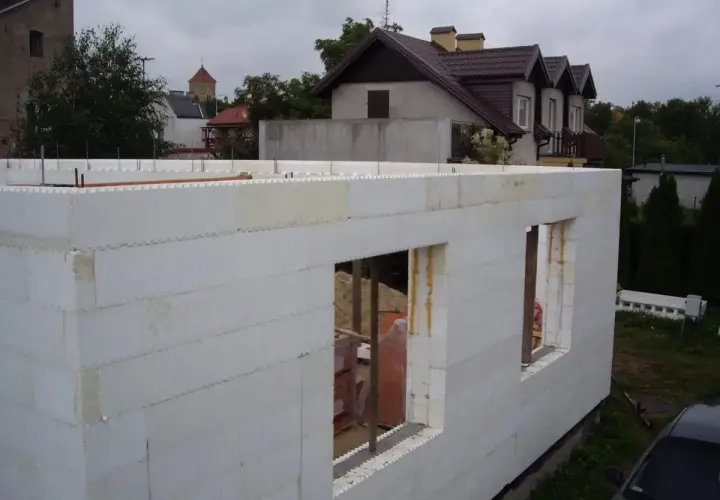 Budowa domu ze styropianu. Białe bloki tworzą ściany z otworami okiennymi. W tle widać inne budynki i drzewa.