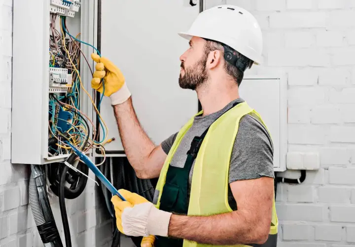 Elektryk w kasku i kamizelce odblaskowej pracuje przy skrzynce z przewodami. Dokładna wycena koszt instalacji elektrycznej wymaga precyzji.