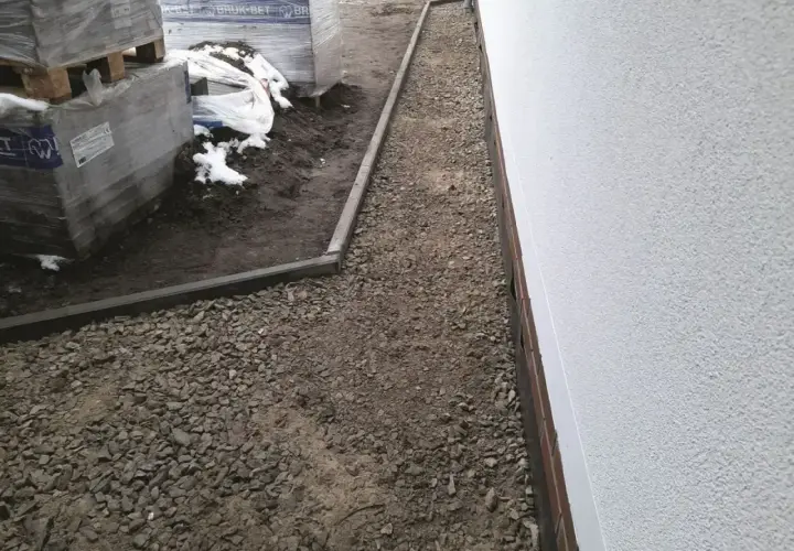Wykonano opaskę wokół domu z kamienia, która chroni fundamenty. Obok leżą palety z materiałami budowlanymi.