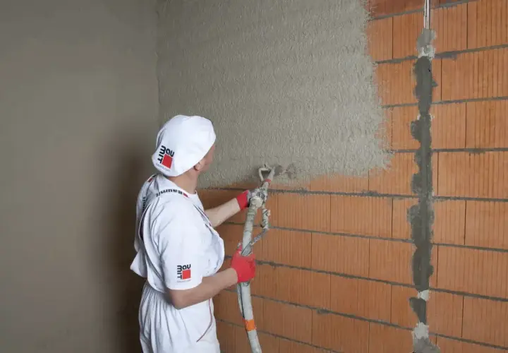 Pracownik w białym stroju nakłada tynki cementowo wapienne na ścianę z pustaków ceramicznych za pomocą agregatu.