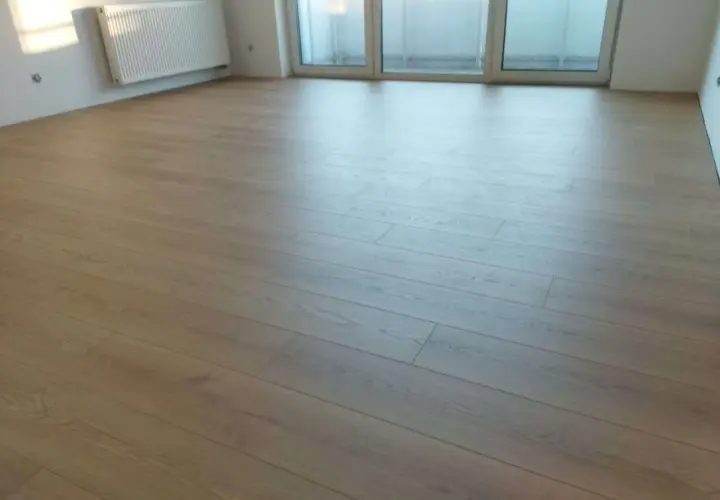 Nowe panele ułożone wzdłuż pomieszczenia, tworząc jasną, drewnianą podłogę. W tle biały grzejnik i okno.