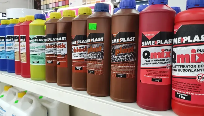 Rzędy butelek z dodatkami do cementu: plastifikatory do betonu i zapraw budowlanych. Różne kolory i pojemności.