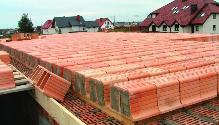 Budowa stropu ackermana. Czerwone pustaki ceramiczne ułożone w rzędach, gotowe do zalania betonem. W tle domy z czerwonymi dachami.