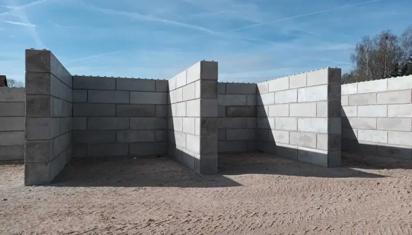 Budowa muru oporowego z bloczków betonowych. Konstrukcja w kształcie litery L, z widocznymi cieniami na piasku.