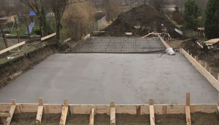 Plac budowy z wylana płytą betonową, przygotowywaną pod fundamenty. Widać zbrojenie i szalunki, wskazujące na różne klasy ekspozycji betonu.