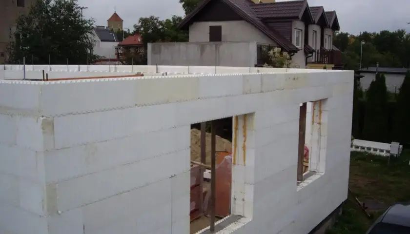 Budowa domu ze styropianu. Białe bloki tworzą ściany z otworami okiennymi. W tle widać inne budynki i drzewa.