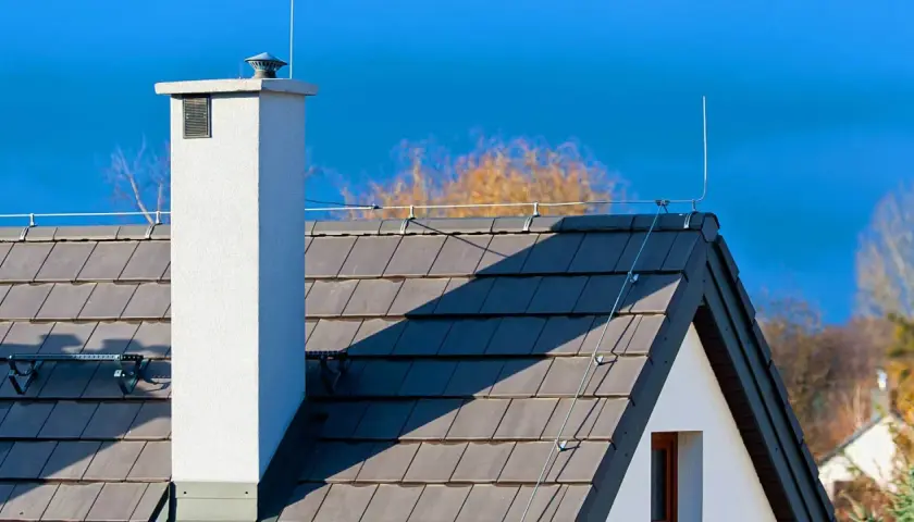 Biały komin i dachówka na tle błękitnego nieba. Na dachu widoczny jest system piorunochronu, chroniący dom przed uderzeniami pioruna.