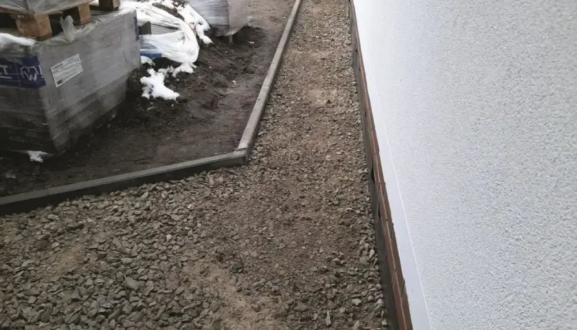Wykonano opaskę wokół domu z kamienia, która chroni fundamenty. Obok leżą palety z materiałami budowlanymi.