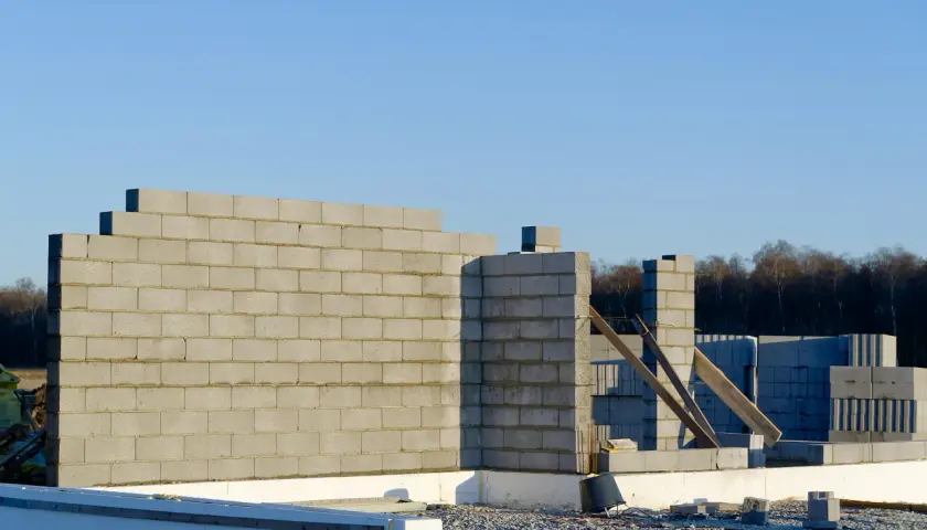 Budowa domu w toku. Wzniesiona ściana nośna z bloczków betonowych, w tle kolejne elementy konstrukcji i las.