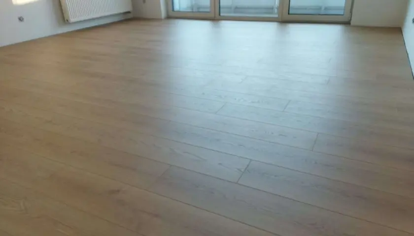 Nowe panele ułożone wzdłuż pomieszczenia, tworząc jasną, drewnianą podłogę. W tle biały grzejnik i okno.