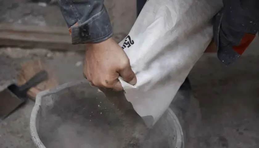 Ręce wsypują cement z worka do wiadra. Różne rodzaje cementu czekają na użycie.
