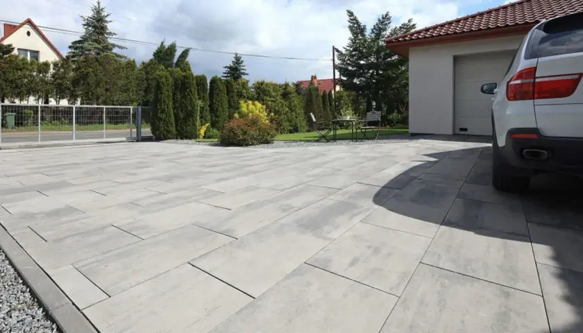 Nowoczesny podjazd z szarej kostki brukowej przed domem z garażem i białym samochodem.