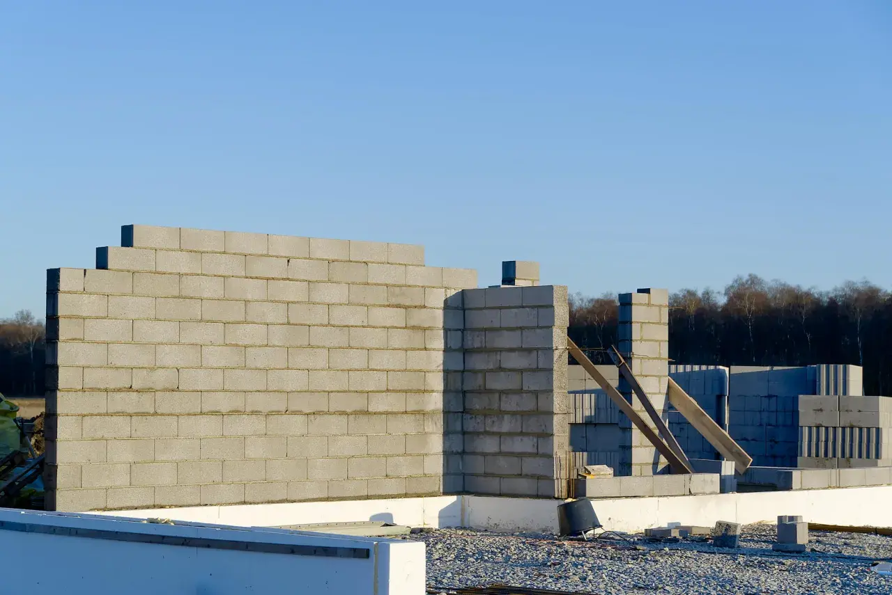 Budowa domu w toku. Wzniesiona ściana nośna z bloczków betonowych, w tle kolejne elementy konstrukcji i las.