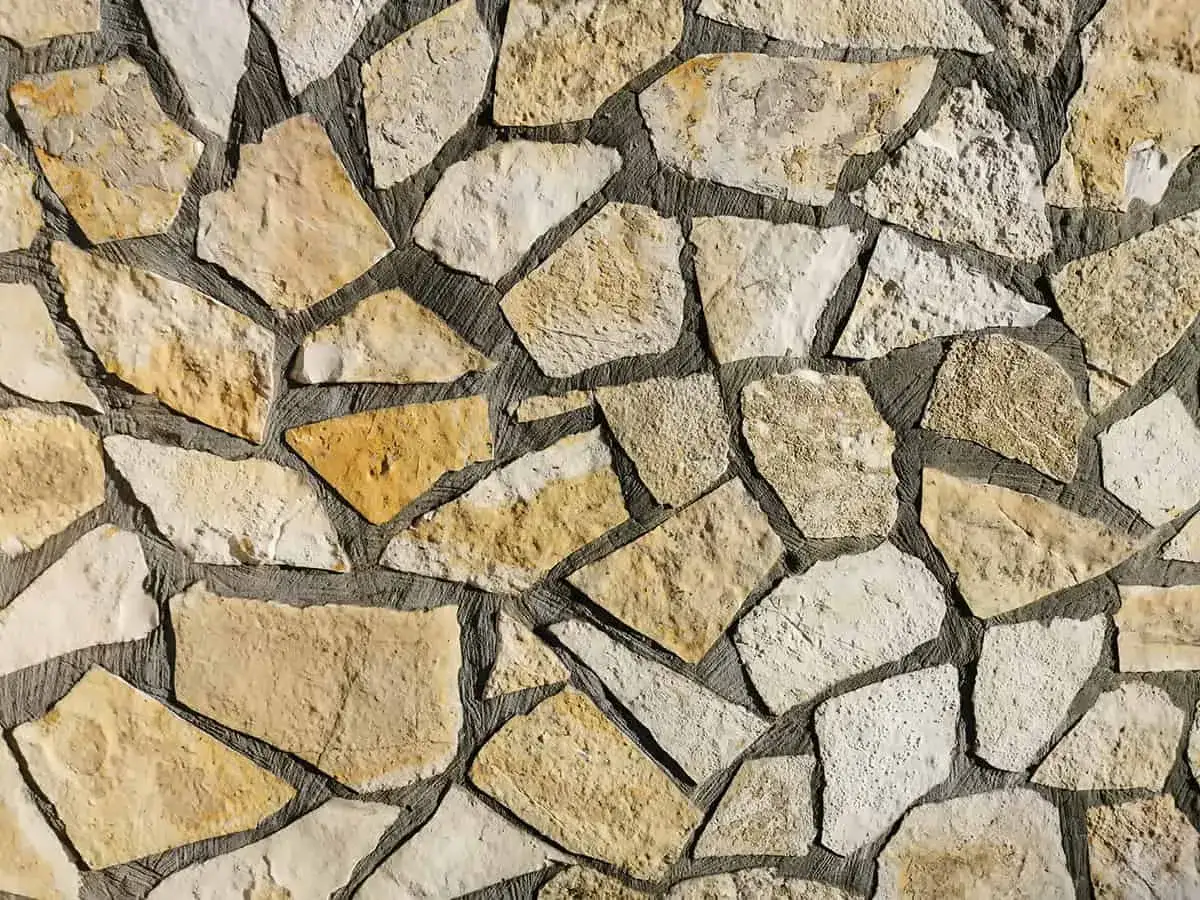 Kamienna ściana z nieregularnych, żółtawych i białych kamieni. Piaskowiec tworzy naturalną, rustykalną mozaikę.