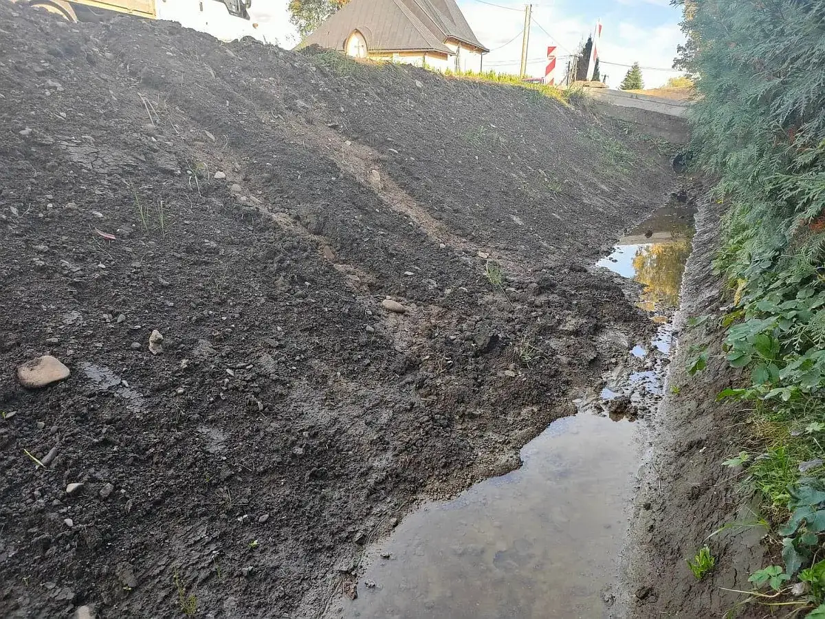 Błotnisty rów melioracyjny z wodą i ziemią po pracach. W tle dom i drzewa.