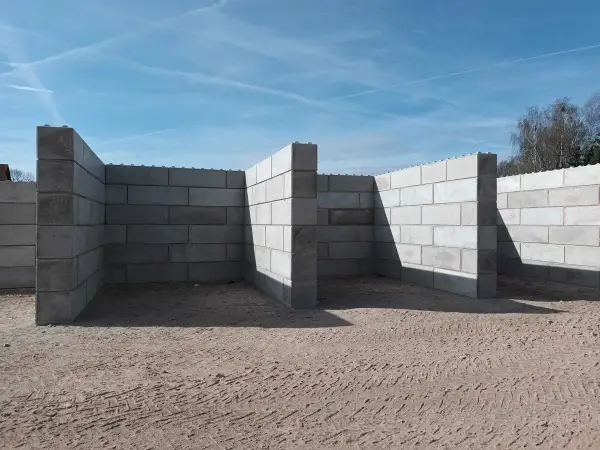 Budowa muru oporowego z bloczków betonowych. Konstrukcja w kształcie litery L, z widocznymi cieniami na piasku.
